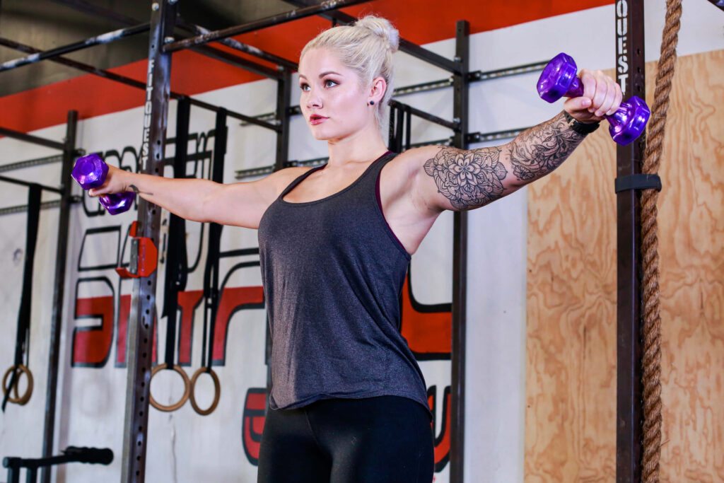 blonde female athlete with sleeve tattoos doing lateral raises with purple jelly bells